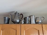 Photo showing six pewter items including a large tea pot, multiple cups and mugs arranged on top of a cabinet.