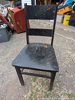 Antique wooden chair painted black, showing paint wear and chipping, photographed outdoors with other household items in the background.