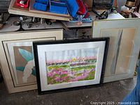 Three framed artworks leaning against a workbench and other items in a workshop space.
