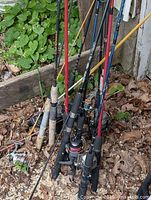 Various fishing rods and reels grouped leaning against a wall and post outdoors on ground with leaves and plants around. Rods have different colors and handle materials.