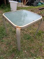 Square metal side table with frosted glass top, visible rust on metal frame and legs, table placed outdoors on grass.