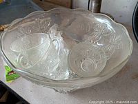 Top-down view of glass punch bowl containing six matching glass cups inside, showing detailed grapevine and leaf pattern on both bowl and cups.