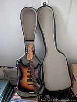 Top-down view of the guitar body and neck inside an open gray hard-shell guitar case showing the sunburst body and the headstock of the neck.