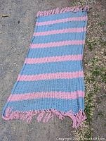 Full view of hand knitted blue and pink striped blanket laid flat on ground showing colors, knit pattern, and fringed edges
