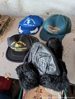 Four hats on a concrete surface: three baseball caps with promotional logos and one black fur-trimmed leather cap.