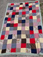 Full view of the large antique patchwork quilt spread out on grass showing various colored and patterned fabric patches sewn in a grid layout along with a floral border edge.