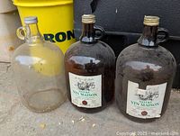 Three large glass wine bottles or jugs, including one clear and two dark brown labeled jugs, placed on ground outdoors.