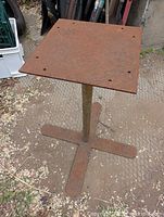 Rusty metal table base with square top plate, showing top surface with pre-drilled holes and cross-legged base on ground.