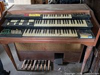 Front view of vintage electric organ synthesizer with two keyboard manuals, control buttons, and foot pedals visible with wooden casing.