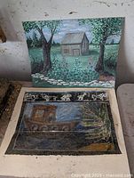 Photo of the pair of paintings, showing the canvas board barn landscape on the wall and the wax painting of Noah's Ark laid on a chair.