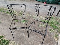 Pair of black iron chairs with decorative backrests and no seats, placed outdoors on gravel ground.