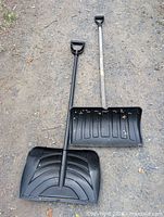 Pair of plastic snow shovels, black scoops and handles, placed on the ground with visible dirt and wear.