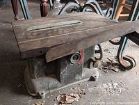 Metal base unit of a portable table top table saw on the floor with leaves and dirt around, showing the top table surface with slot and adjustment handles on the side.