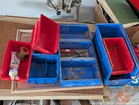 Red and blue plastic tool boxes arranged on a flat surface. Boxes vary in size and shape with some compartments and dividers visible. Surfaces show some dirt and wear.