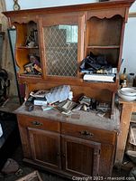 Front view of entire vintage maple china hutch showing upper shelving with mirrored door and lattice, lower cabinet with two doors and drawers. Hutch surfaces cluttered and dusty, wood finish medium-brown with scalloped top edge.