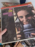 Close up of one vinyl album cover titled 'Auf Zum Schützen Fest,' featuring a person smiling and holding a metallic object. Shows condition with wear and creasing.