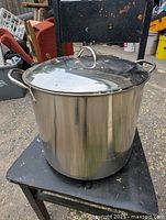 Front view of an 18-inch tall stainless steel stock pot with lid, placed on a black wooden chair outdoors; pot has two side handles and a lid handle, shows usage marks.