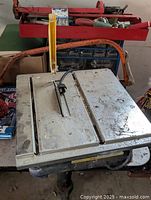 Front angled view of electric tile cutting saw showing metal deck, circular saw blade with yellow guard, and orange power cord wrapped around base.