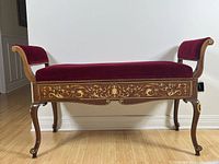 Front angled view of antique wooden bench with floral inlay and red velvet upholstery on seat and scroll arms