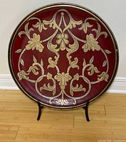 Front view of large round red and cream carved wooden decorative plate with floral design, displayed upright on black stand on wooden floor.
