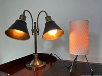 Photo shows a brass double-headed desk lamp with black shades lit, beside a beige textured lamp also lit on tripod legs.