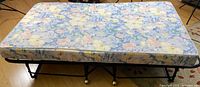Full view of single fold up cot bed with metal frame and floral mattress on a hardwood floor.