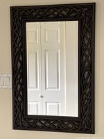 Full view of the rectangular wall-mounted hall mirror showing the walnut coloured resin frame with an intricate woven design and the mirror surface reflecting a door.