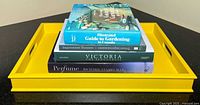 Yellow wooden decor tray with five hardcover books stacked one on top of another, titles clearly visible from top down.