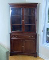 Full front view of the mahogany corner cabinet showing glass doors on upper portion and wooden doors below with drawer in between.