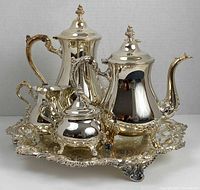 Front view of silver plated coffee and tea set on ornate tray, showing coffee pot, teapot, creamer, sugar bowl with broken handle and foot.