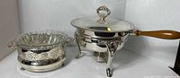 Silver plate serving stand with a removable glass insert and a silver plated chafing dish with a wooden handle shown side-by-side on a white background.