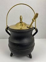 Black cast iron smudge pot with brass lid and handle shown from front angle with brass fire starter wand in lid slot.
