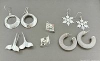Lot of 5 pairs of sterling silver pierced earrings arranged on gray background including shaped hoops, snowflakes, textured hoops, sculptural dangles, and small studs.