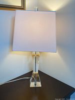 Full view of table lamp showing clear acrylic base and off-white rectangular shade on a dark table.