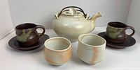 Full view of teapot in center with two matching ceramic cups and saucers to left and right and two smaller off-white tea ceremony cups in front
