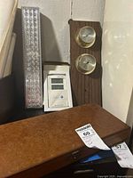 Photo showing Bell and Howell light, digital thermometer, wooden barometer and thermometer mounted on wood, and antique print set closed box.