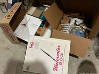 Boxes containing watercolor blocks, brushes, palette, carving tools, and art supplies.