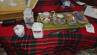 Lot display showing Asian themed china pieces on a red plaid cloth, including a Rose Mandarin demi cup and saucer, porcelain sugar bowl with spoon, small hand-painted dishes, and metal utensil rests.