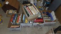 Five cardboard boxes filled with assorted paperback and hardcover books of various sizes and colors. Boxes are set on a concrete floor near paint cans and garage items.
