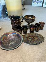 Full set displayed on floor showing pitcher, six cups, compote, bowl, and oval dish with grape and floral patterns in iridescent carnival glass.
