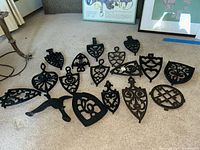 Overview photo showing all 16 cast iron trivets in a group on carpet, various shapes and sizes including geometric and ornate shield shapes.