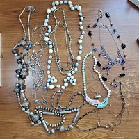 Overview photo showing multiple necklaces laid flat on wood surface with various bead types and colors including transparent, blue, white and silver beads.