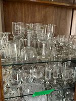 First photo showing multiple clear glass stemware on shelves including wine glasses, cocktail glasses, and cordial glasses with varied shapes and sizes.