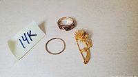 Three yellow gold jewelry pieces: multi-stone ring, plain hoop earring, and floral brooch pin displayed with 14k marker card