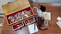 View of open vintage lacquered wood jewelry box filled with various jewelry beads and pieces, plus watch and belt buckle on surface.