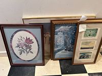 Four framed prints leaning against a wall on a checkered floor, showing floral, landscape, and architectural images.