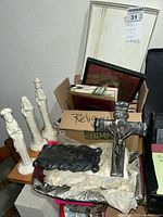 Photo showing white ceramic figurines with distinct headwear and robes, placed on a wooden chair next to a black decorative cross and some framed artworks in a box labeled 'religious'.