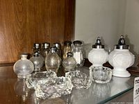 Photo showing multiple glass salt and pepper shakers, including two white milk glass with metal tops, and four small clear glass dishes in front.