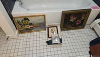 Four framed wall art pieces displayed on a tiled floor near bathtub, including two larger paintings and two smaller framed prints inside a box.