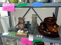 Photo showing white hobnail milk glass vase, clear glass vase, brown artisan pottery vase, beach stones, miniature Mexican pots, and wooden salad bowls stacked together on shelf.